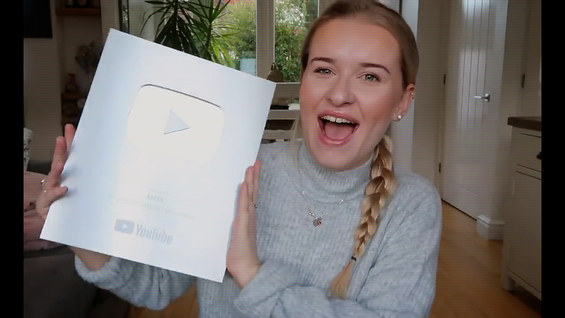 Jazzy Whipps is smiling to the camera with her blonde hear in a braid, wearing a light grey turtleneck jumper. She is holding a silver YouTube Creator Award to mark reaching 100,000 followers.